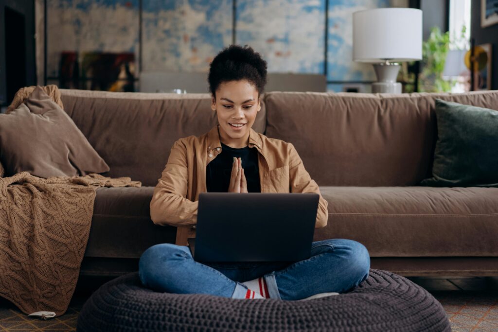 A woman sitting on the floor in her living room in front of her couch and working on her laptop.