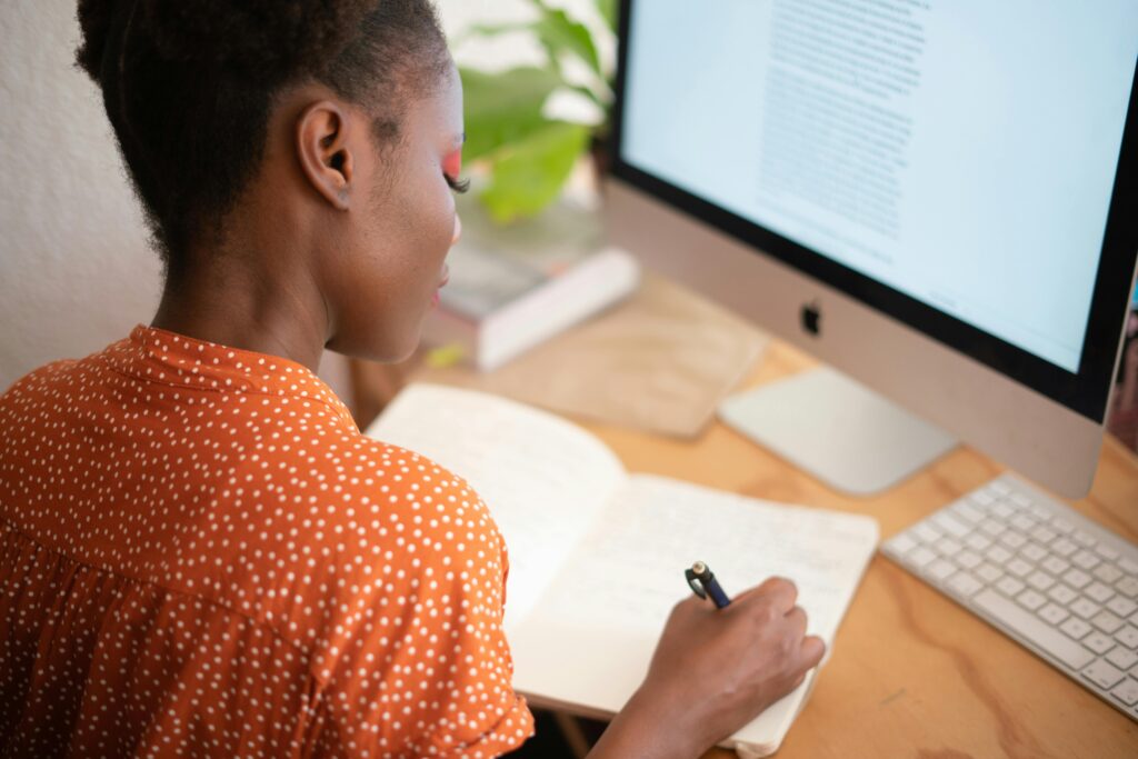 A girl in an orange shirt writing in her notebook and sitting behind a computer screent.